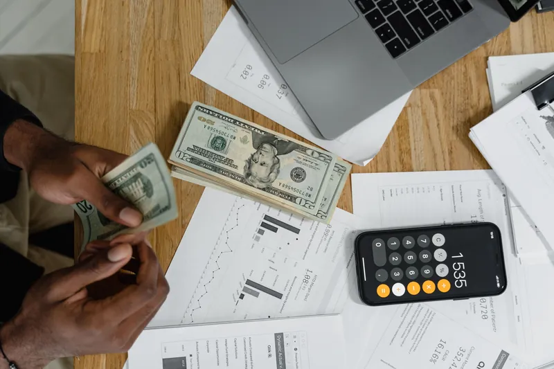 Des mains comptent des billets au-dessus de relevés chiffrés et de graphiques, à côté d'une calculatrice de smartphone et d'un ordinateur portable